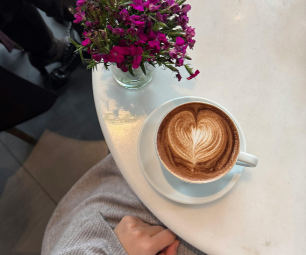 Na sliki je miza s skodelico kave in vazo vijoličnih rož